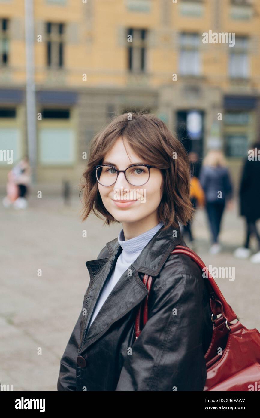 Portrait of young woman in glasses and leather cloak in middle of city Stock Photo