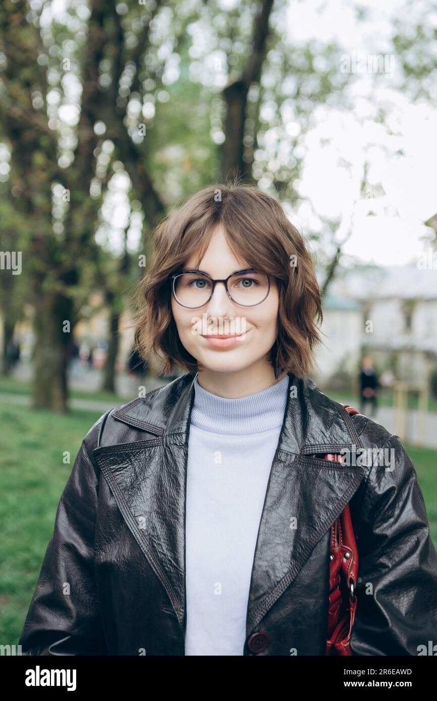 Portrait of young woman in glasses and leather cloak in middle of city Stock Photo