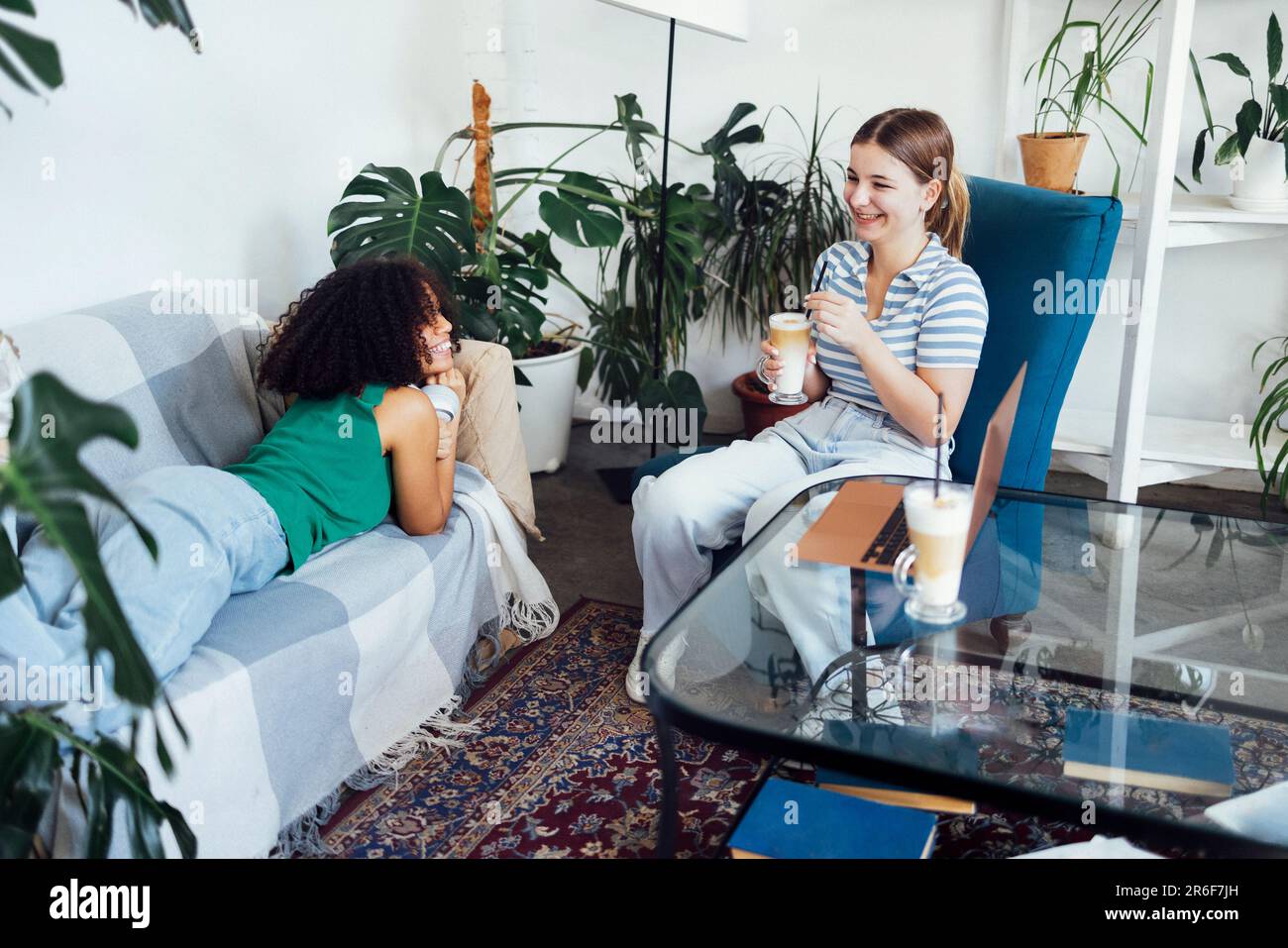 Cute multiracial teenagers drink cappuccino and chat. Teen girls have good time together and watch movie on laptop. Smiling female friends having fun Stock Photo