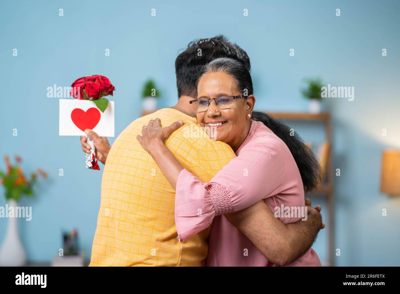 Happy indian adult son hugging or embracing to senior mother by giving red roses with greeting card at home - concept of mother's day, relationships Stock Photo