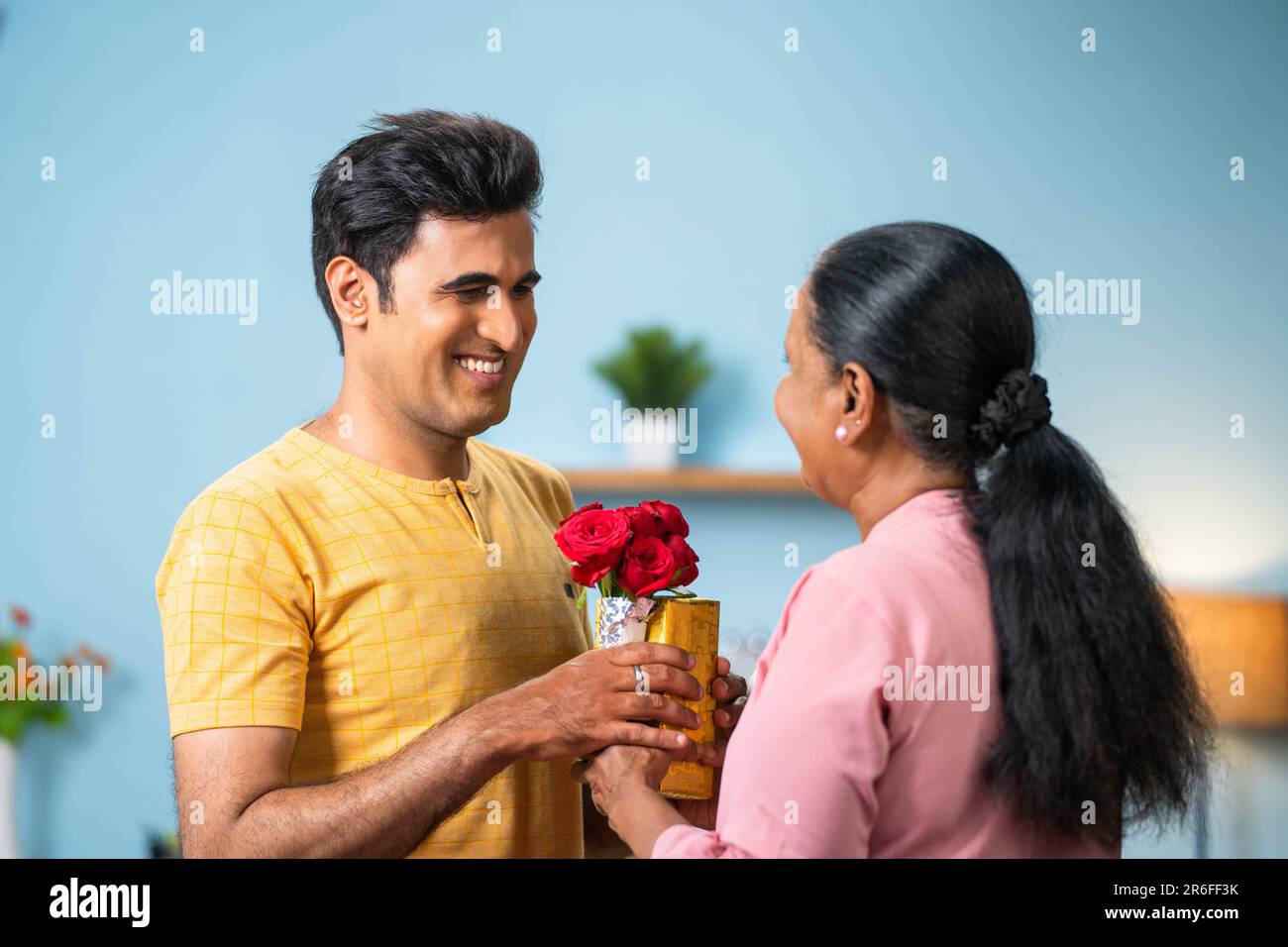 Indian adult son giving flowers with gift to senoir mother during mother day celebration - concept of relationship bonding, birthday present and happy Stock Photo