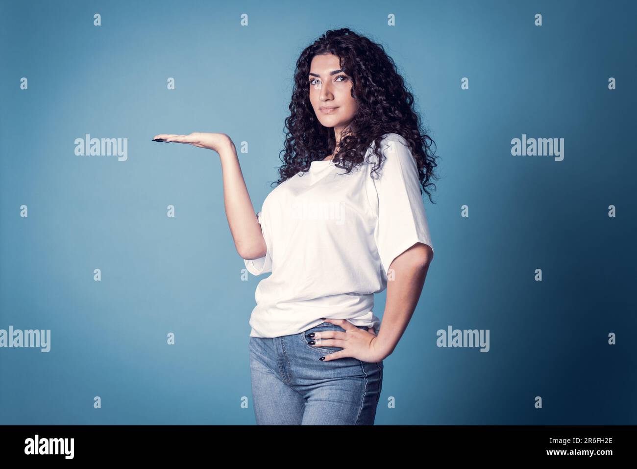 Shot of serious confident curly woman, wears casual basic solid white t-shirt holding isolated over blue background. Stock Photo