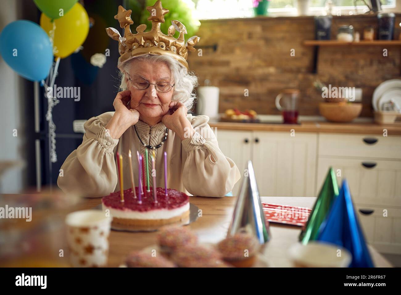 Elderly elegant gray haired senior woman wearing a golden crown celebrating her birthday, sitting in front of birthday cake. Home, lifestyle, holiday Stock Photo