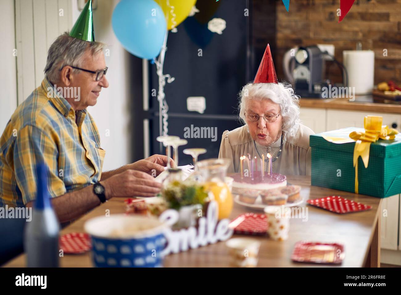 Happy senior woman blowing candles on her birthday cake, sitting at table with husband cheering her. Celebration, lifestyle, senior life concept. Stock Photo