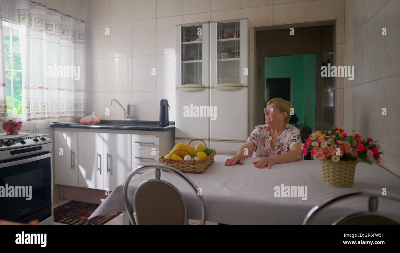 Contemplative senior woman sitting at kitchen table looking out window ...