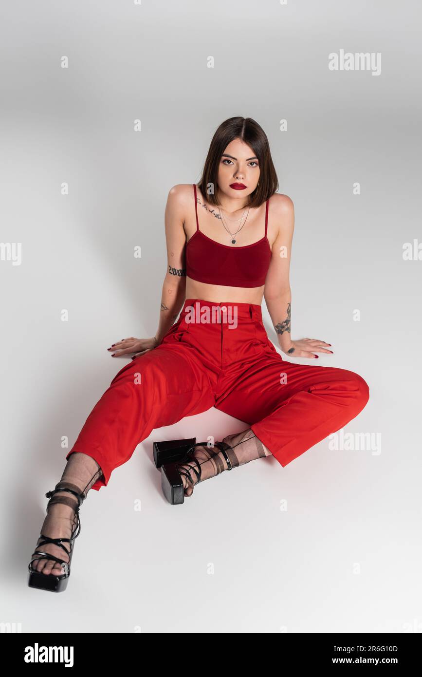 fashion trend, young model in red outfit, tattooed woman with short hair and nose piercing posing in red crop top and pants on grey background, genera Stock Photo
