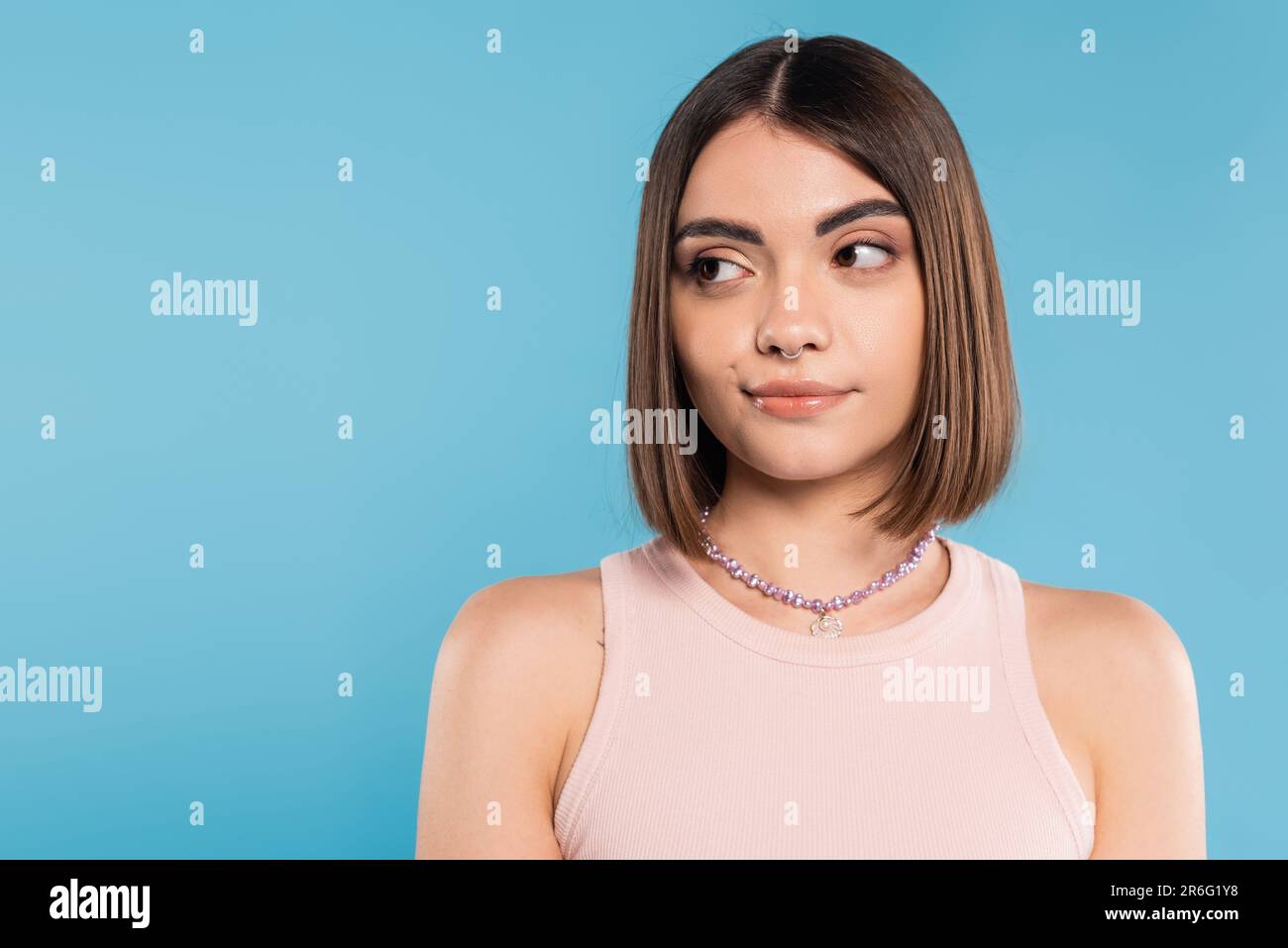 doubting, young brunette woman with short hair and nose piercing standing in tank top and looking away on blue background, youth culture, skepticism, Stock Photo