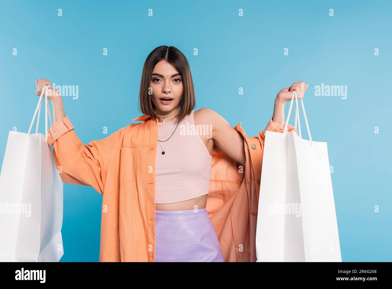 summer trends, pierced young woman in fashionable outfit posing with shopping bags on blue background, casual attire, stylish, generation z, modern fa Stock Photo