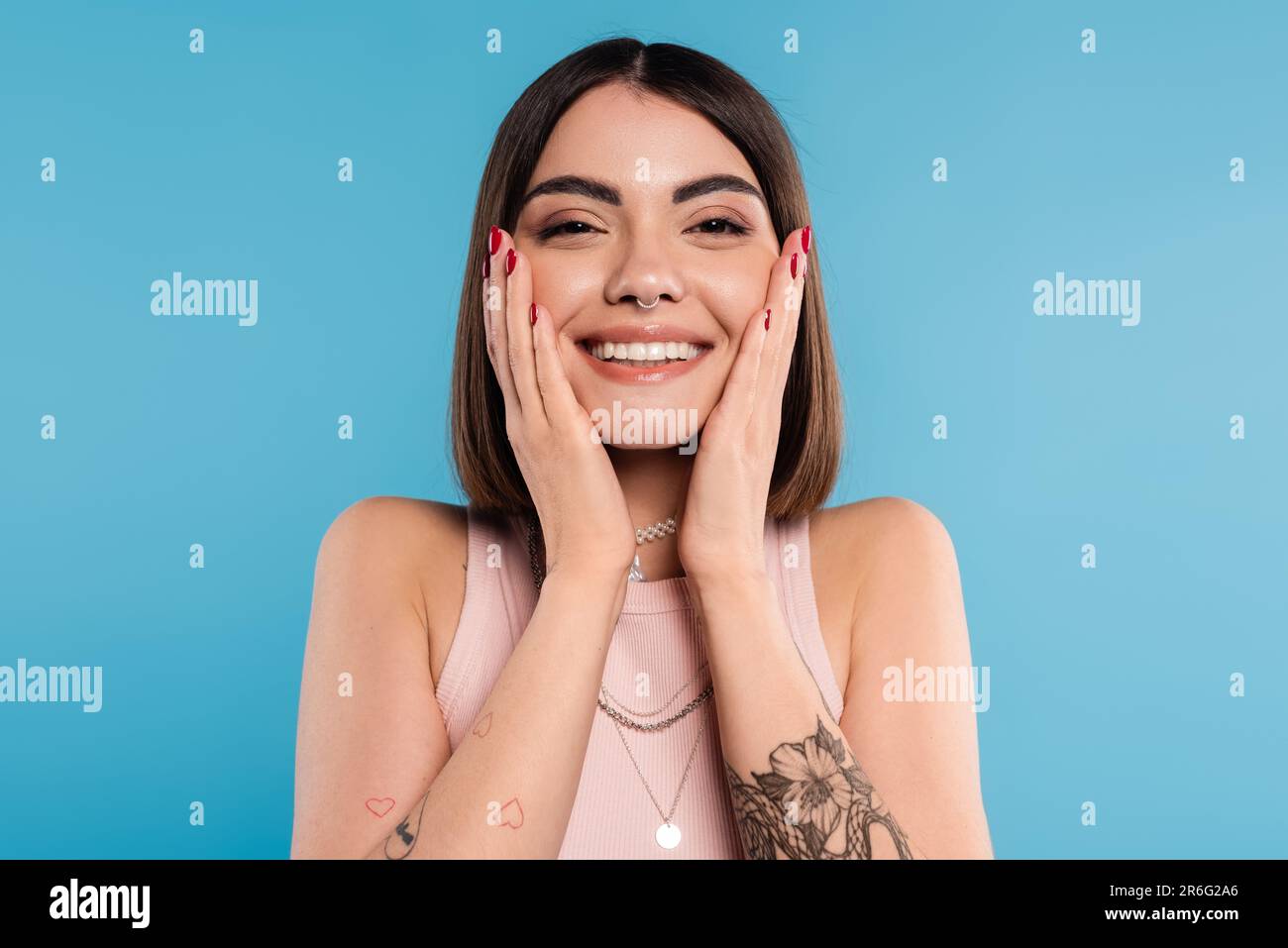 positivity, tattooed young woman with short brunette hair in tank top smiling  and touching her face on blue background, casual attire, gen z fashion, Stock Photo