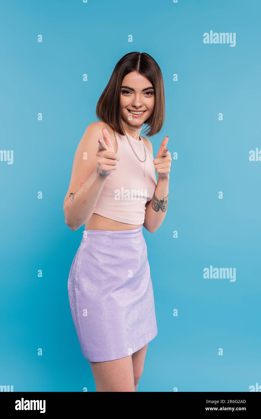 positivity, tattooed young woman with short hair in tank top and skirt smiling and pointing at camera on blue background, casual attire, gen z fashion Stock Photo