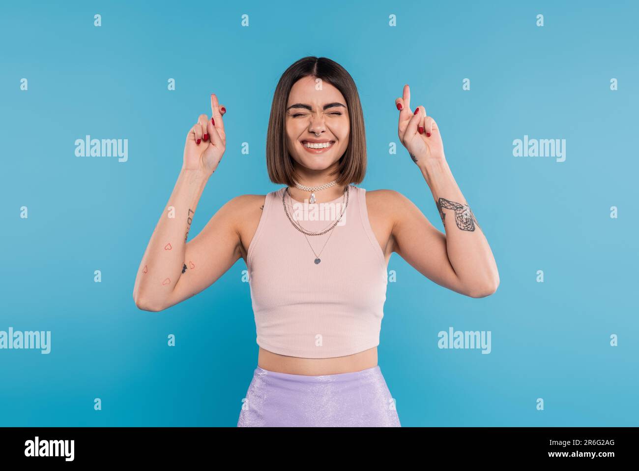 fingers crossed tattooed young woman with short brunette hair in tank top making wish on blue background casual attire gen z fashion happiness sm Stock Photo Alamy