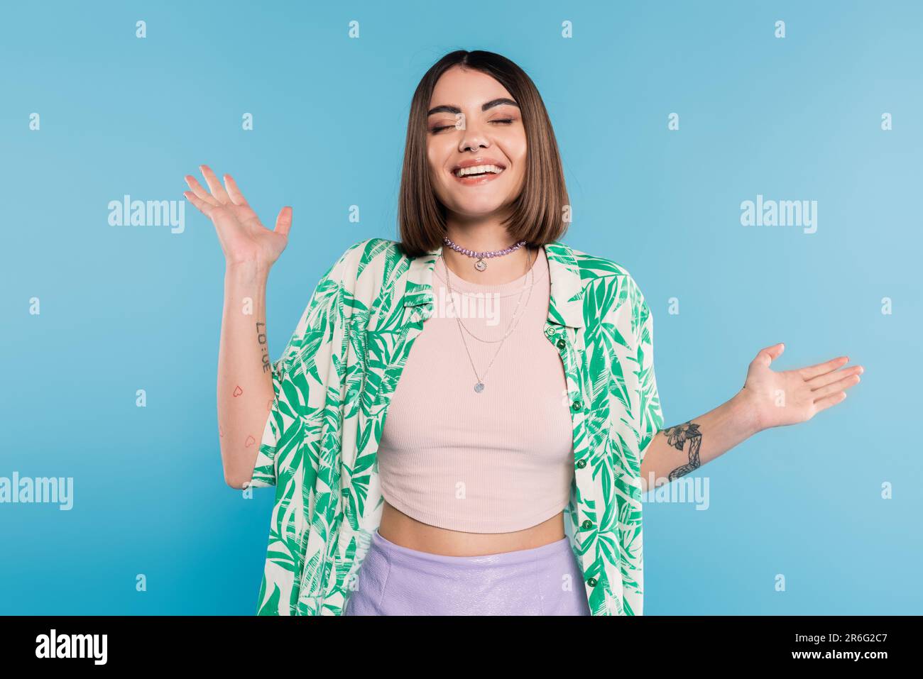 tattooed young woman with short brunette hair wearing shirt with palm tree print, smiling with closed eyes and gesturing with hands on blue background Stock Photo