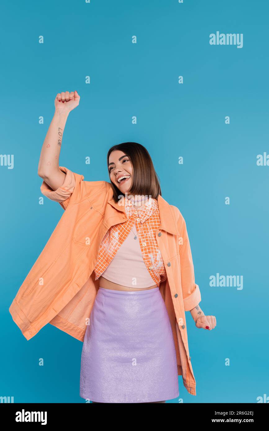 summer outfit, excited young woman smiling while gesturing on blue background, summer outfit, generation z, short brunette hair, orange shirt, pierced Stock Photo