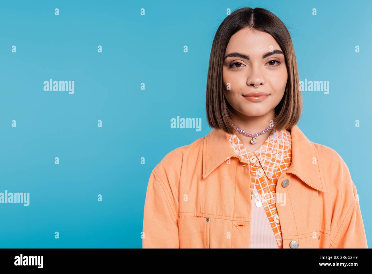 generation z, portrait of pretty woman, young fashion model looking at camera on blue background, orange shirt, short brunette hair, pierced nose, sum Stock Photo