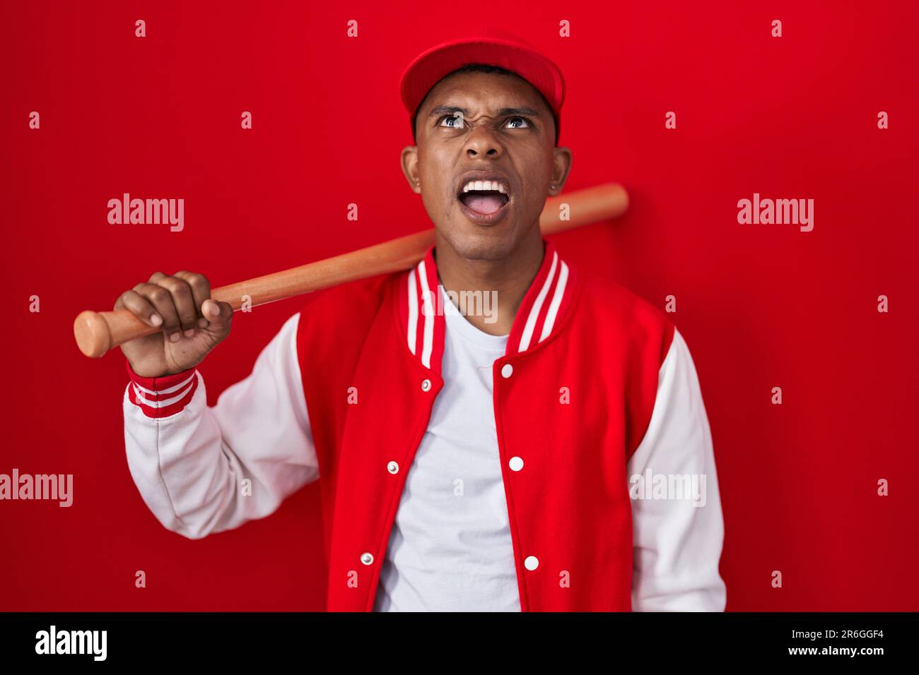 Young hispanic man playing baseball holding bat angry and mad screaming ...
