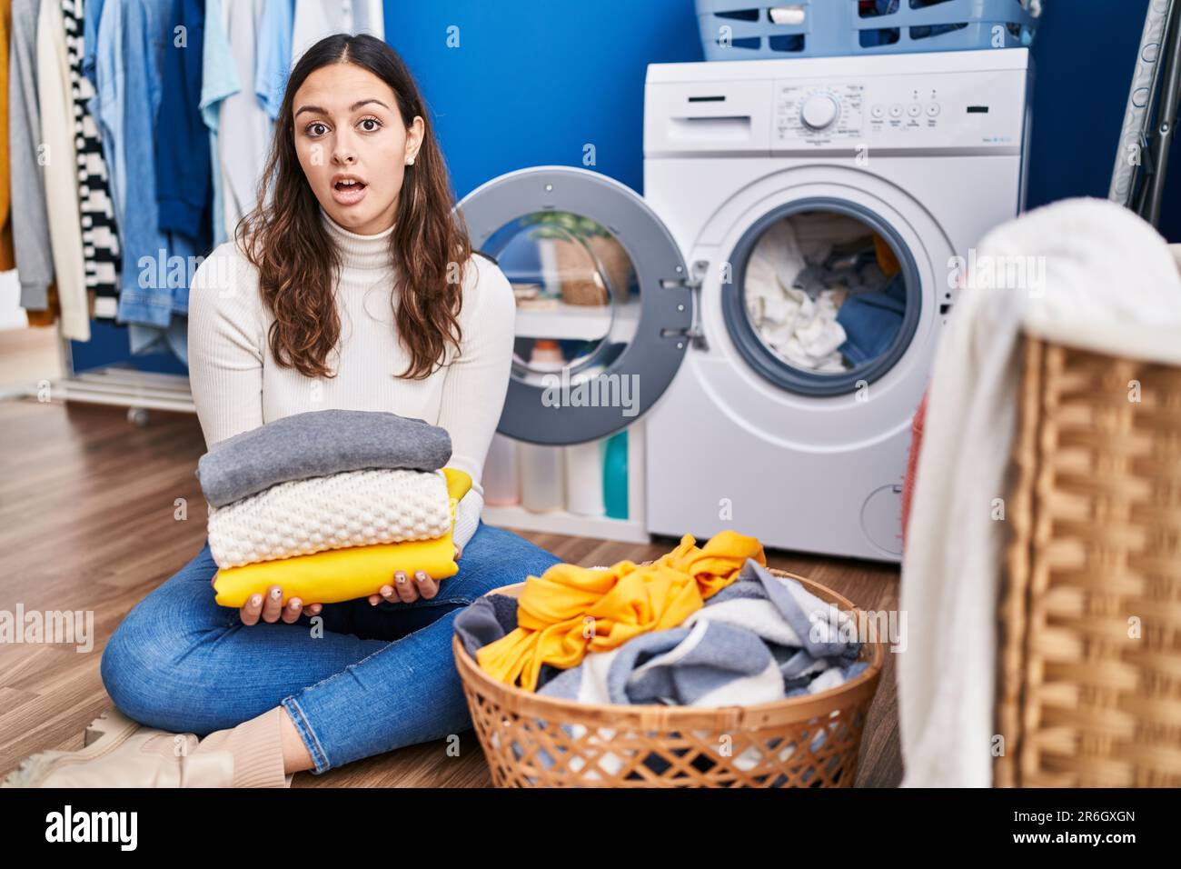 Young hispanic woman holding clean laundry in shock face, looking