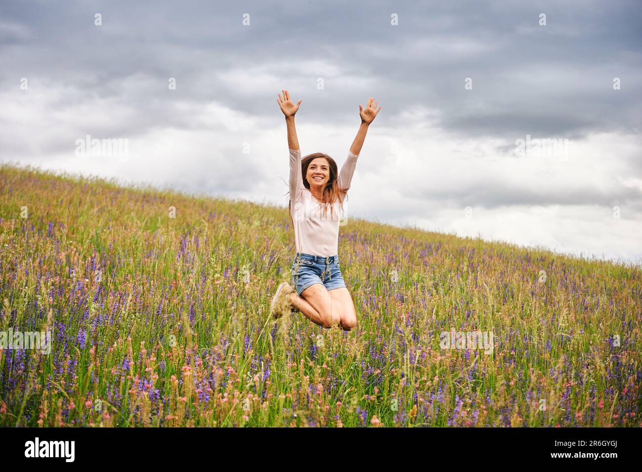 Young beautiful woman hiking in field with wildflowers, excited girl jumping high Stock Photo