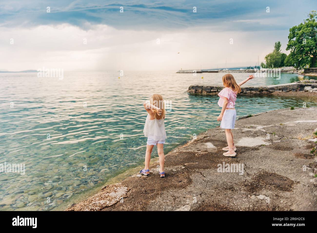 Little girls playing by the lake in summertime, children having fun outside, throwing stones to water, back view Stock Photo