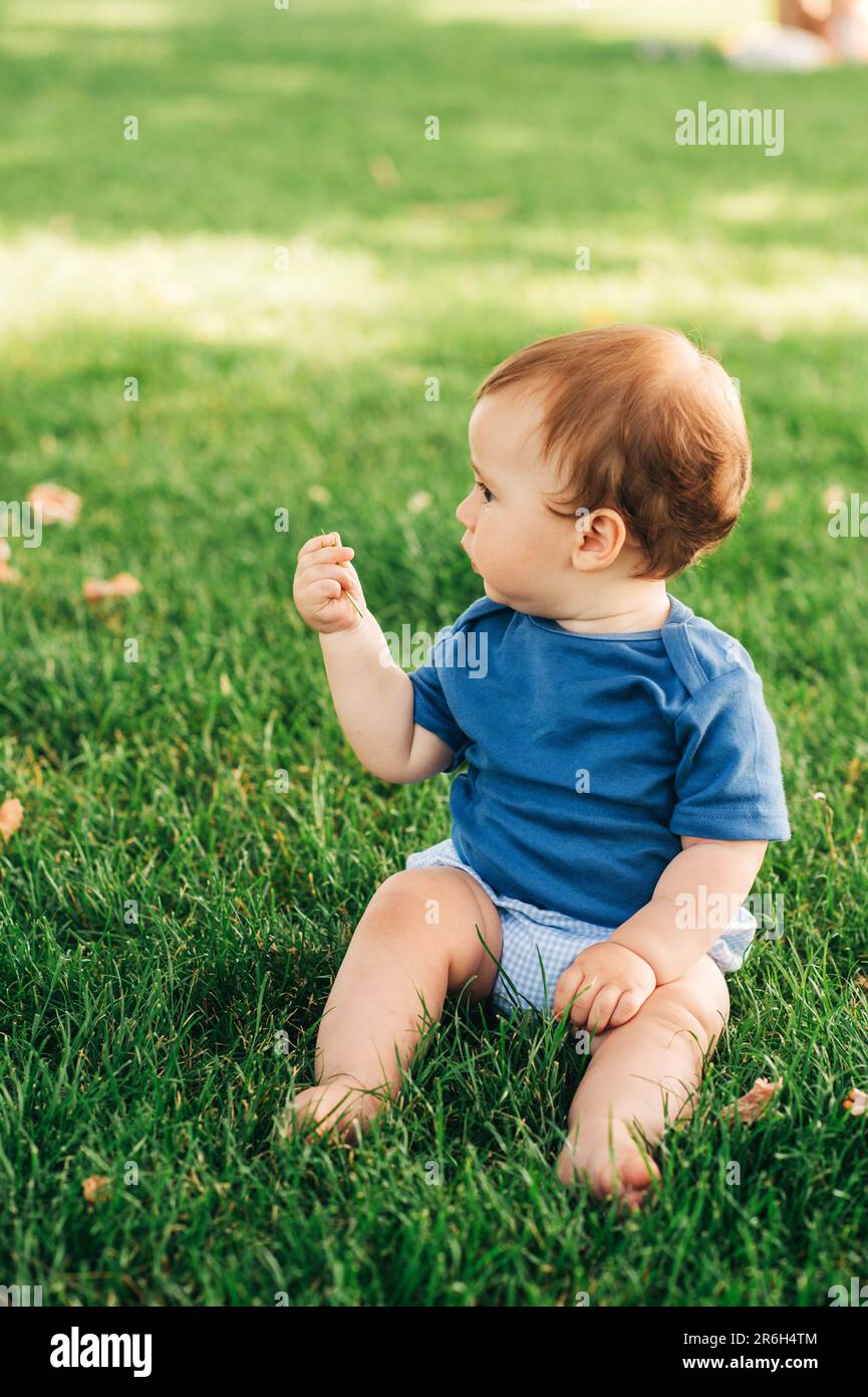 Outdoor portrait of adorable redheaded baby boy playing in summer parc, sitting on green grass, wearing blue bodysuit Stock Photo