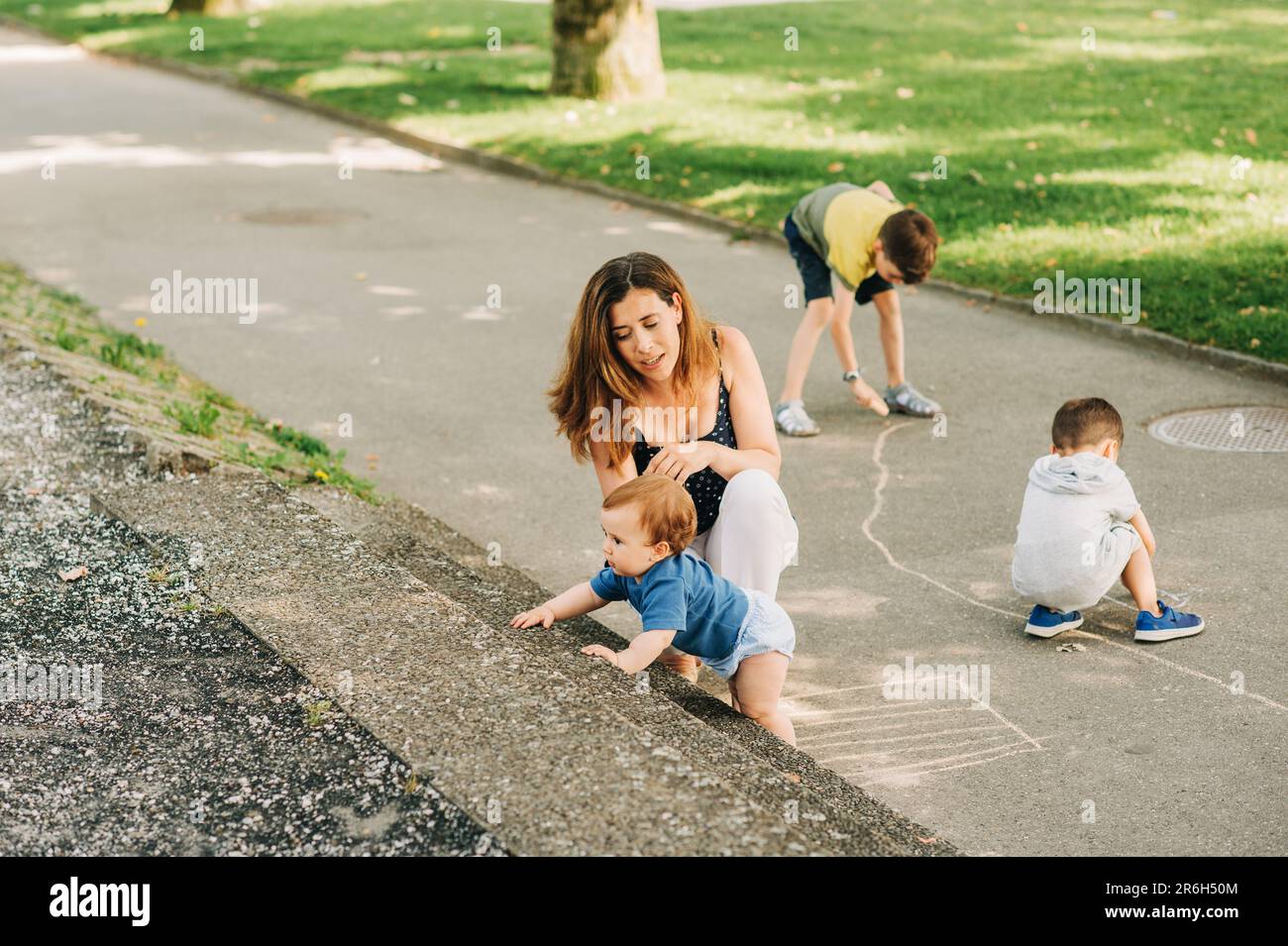 Happy mother spending time with children in summer park, playing with baby, boys drawing with chalk Stock Photo