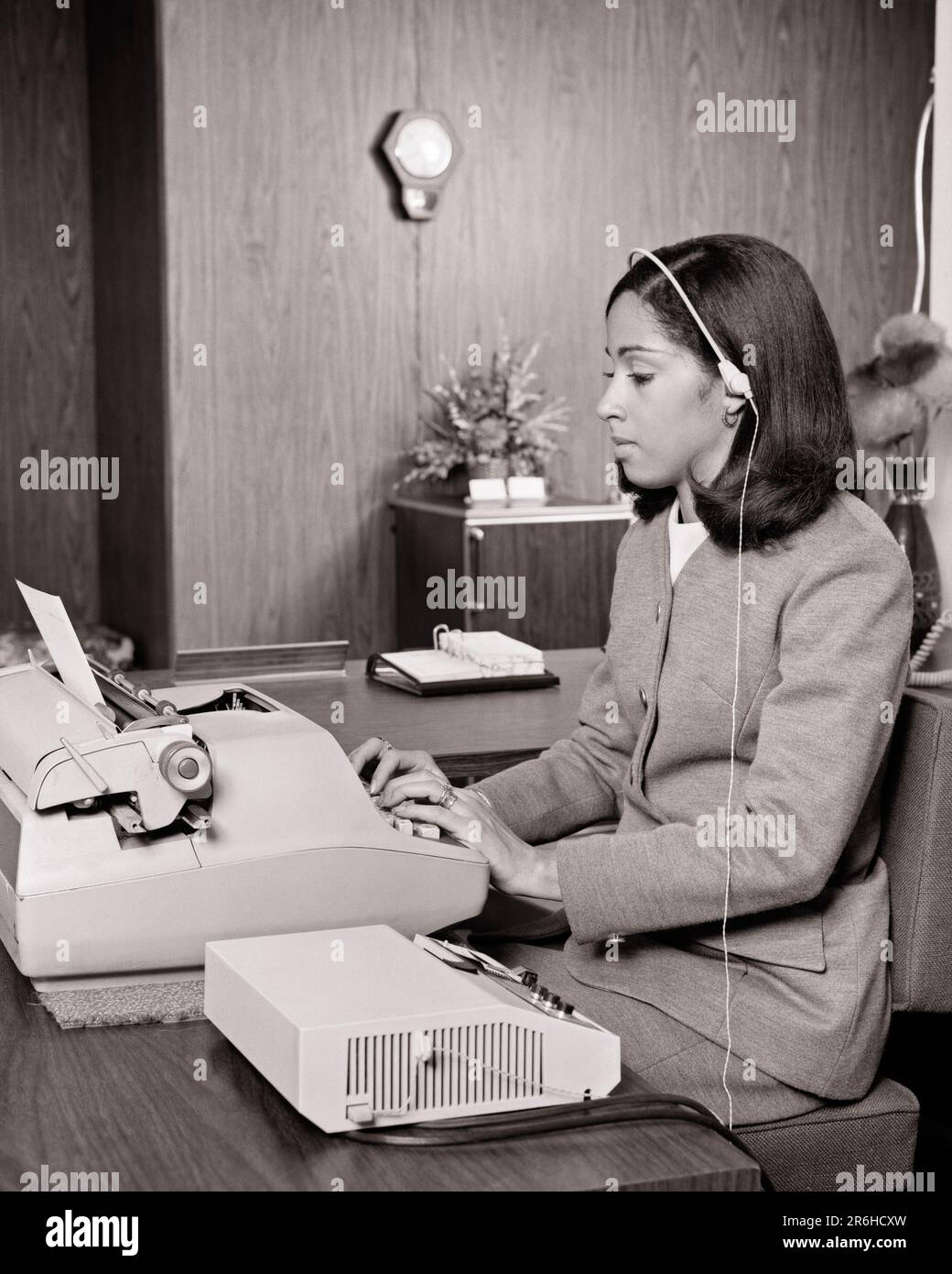 1970s AFRICAN-AMERICAN WOMAN IN OFFICE TYPING ON ELECTRIC TYPEWRITER LISTENING TO DICTATION FROM DICTAPHONE WITH HEADSET - o3035 HAR001 HARS HALF-LENGTH LADIES PERSONS CONFIDENCE B&W HEADSET DICTAPHONE SKILL OCCUPATION SKILLS TYPING AFRICAN-AMERICANS AFRICAN-AMERICAN BLACK ETHNICITY OFFICE WORKER OCCUPATIONS GAL FRIDAY ADMINISTRATOR LISTING SECRETARIES STYLISH SUPPORT AMANUENSIS DICTATION RECORDED YOUNG ADULT WOMAN BLACK AND WHITE CLERICAL HAR001 OLD FASHIONED TYPIST AFRICAN AMERICANS Stock Photo