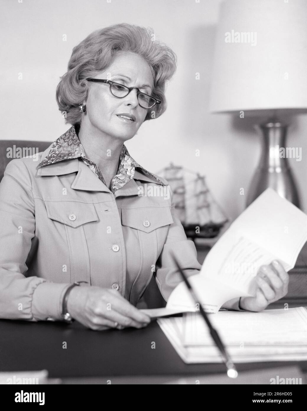 1970s MATURE WOMAN IN OFFICE SITTING AT DESK READING A DOCUMENT WEARING A BUSINESS SUIT AND HALF RIM READING GLASSES - o4076 HAR001 HARS SATISFACTION FEMALES JOBS MANAGER COPY SPACE HALF-LENGTH LADIES PERSONS PROFESSION CONFIDENCE EXECUTIVES MIDDLE-AGED B&W SUCCESS SKILL OCCUPATION SKILLS MIDDLE-AGED WOMAN DOCUMENT AND CAREERS KNOWLEDGE LEADERSHIP HALF AUTHORITY OCCUPATIONS BOSSES STYLISH ATTORNEY MANAGERS RIM BLACK AND WHITE CAUCASIAN ETHNICITY HAR001 OLD FASHIONED Stock Photo