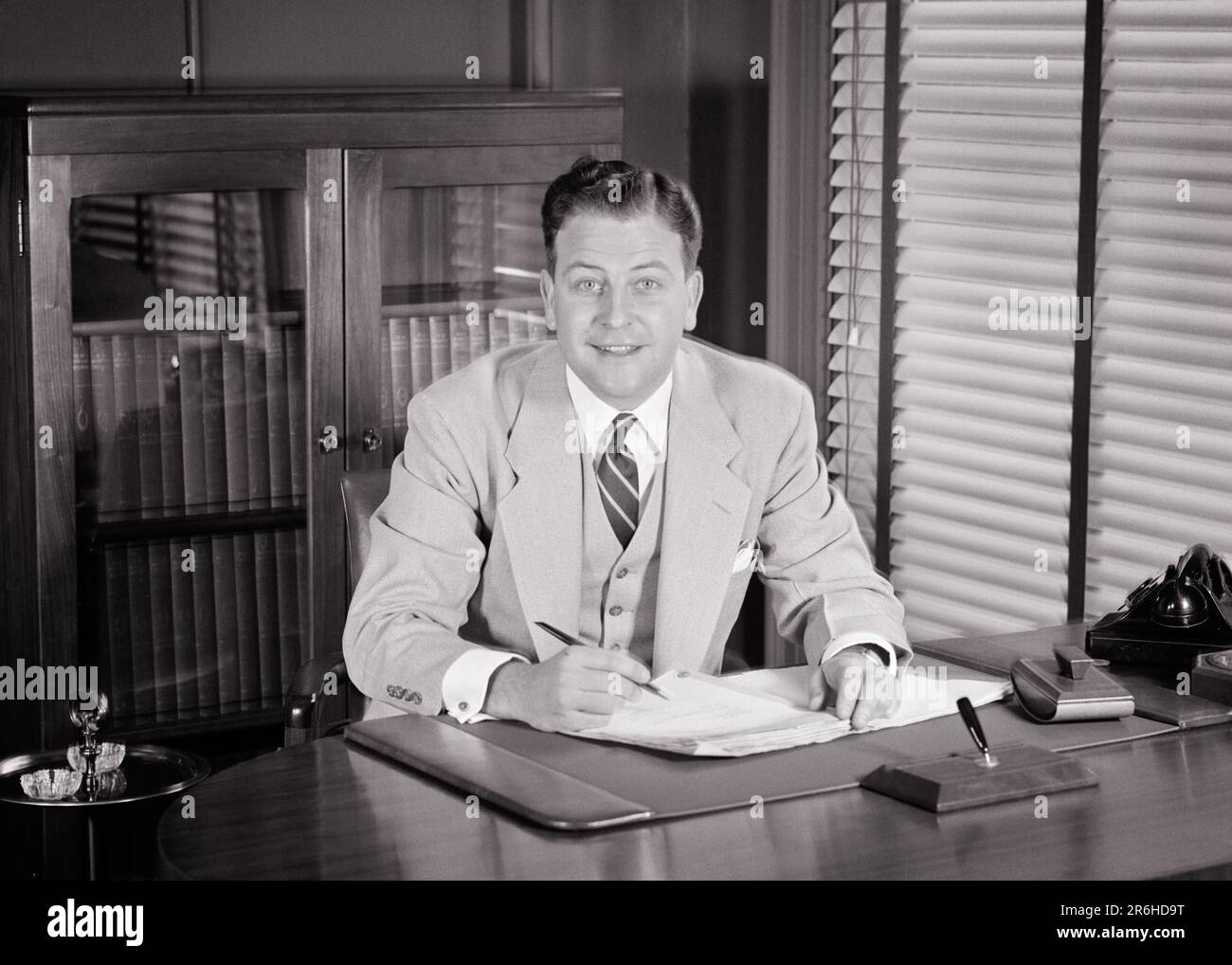 1950s SMILING BUSINESS MAN LOOKING AT CAMERA WEARING THREE PIECE SUIT SITTING AT DESK SIGNING PAPERS - o635 HAR001 HARS COMMUNICATION SIGNING INFORMATION PIECE PLEASED JOY LIFESTYLE SATISFACTION JOBS STUDIO SHOT MANAGER COPY SPACE HALF-LENGTH PERSONS MALES CONFIDENCE EXECUTIVES VEST B&W WIDE EYE CONTACT GOALS SKILL BUG-EYED SUIT AND TIE OCCUPATION HAPPINESS SKILLS CHEERFUL STRATEGY LEADERSHIP PRIDE OPPORTUNITY OCCUPATIONS SMILES BOSSES THREE PIECE SUIT JOYFUL STYLISH BLOTTER WIDE-EYED ATTORNEY MANAGERS MID-ADULT MID-ADULT MAN STARTLED VENETIAN BLINDS BLACK AND WHITE CAUCASIAN ETHNICITY Stock Photo