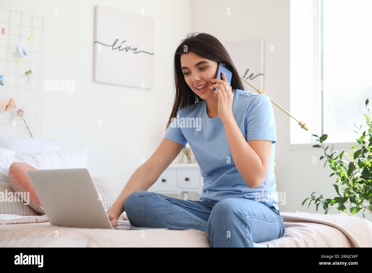 Female freelancer with laptop talking by mobile phone in bedroom Stock Photo