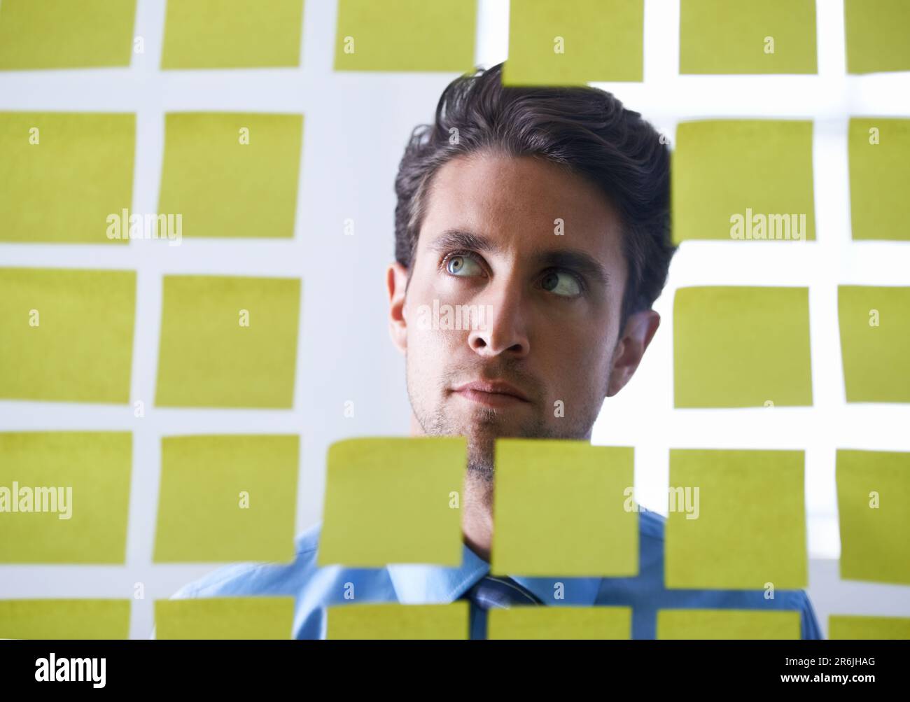Creative man, face and thinking for sticky note, brainstorming or planning tasks on glass board at office. Male person in problem solving, idea Stock Photo