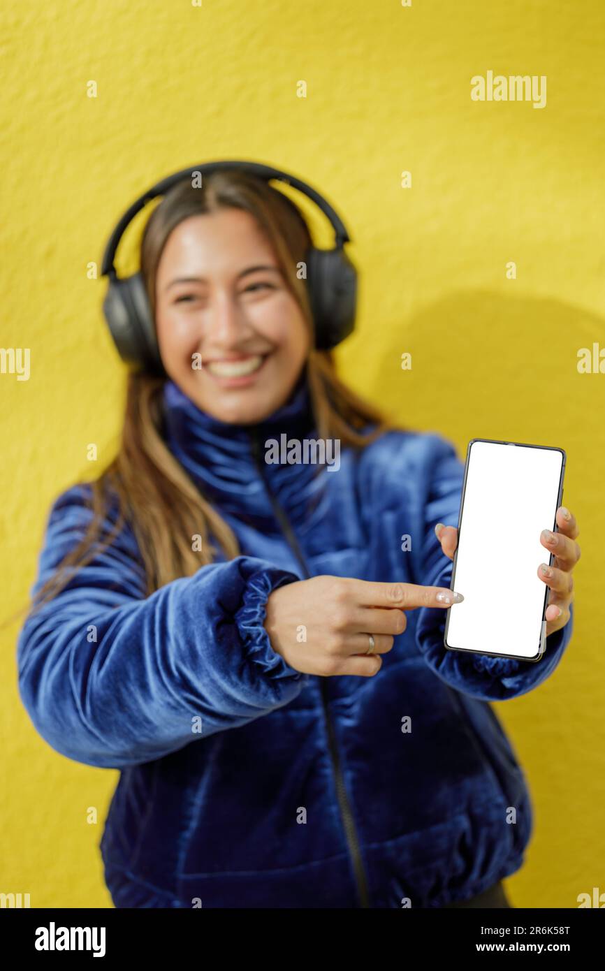 Smiling latin girl with headphones out of focus shows the blank screen of her mobile phone. Stock Photo