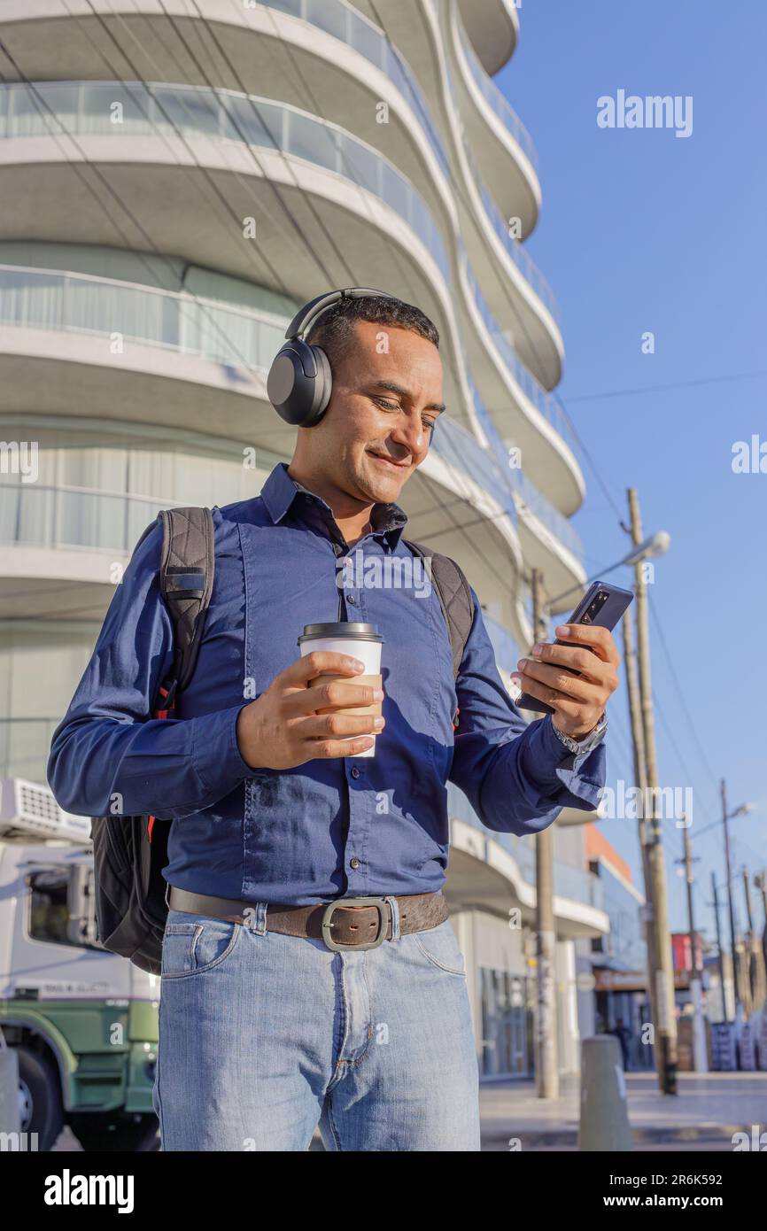 Young latin man with headphones looking at his mobile phone and holding a paper cup with coffee in his hand. Stock Photo