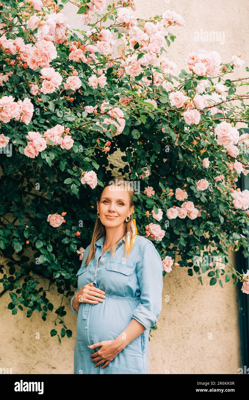 Outdoor portrait of stylish pregnant woman posing in rose garden, wearing denim dress Stock Photo
