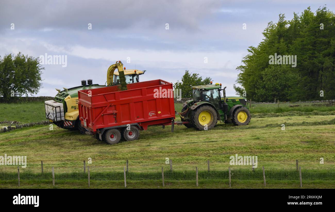 John Deere 6155R tractor haymaking working in scenic farm pasture field (filling loading pulled wagon, cut grass silage, farmer driving) - England UK. Stock Photo