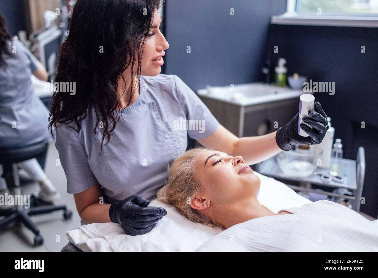 Pretty caucasian beautician communicates with her female client. Beauty master holds cosmetics in her hands and shows them to blod woman. Apparatus fo Stock Photo