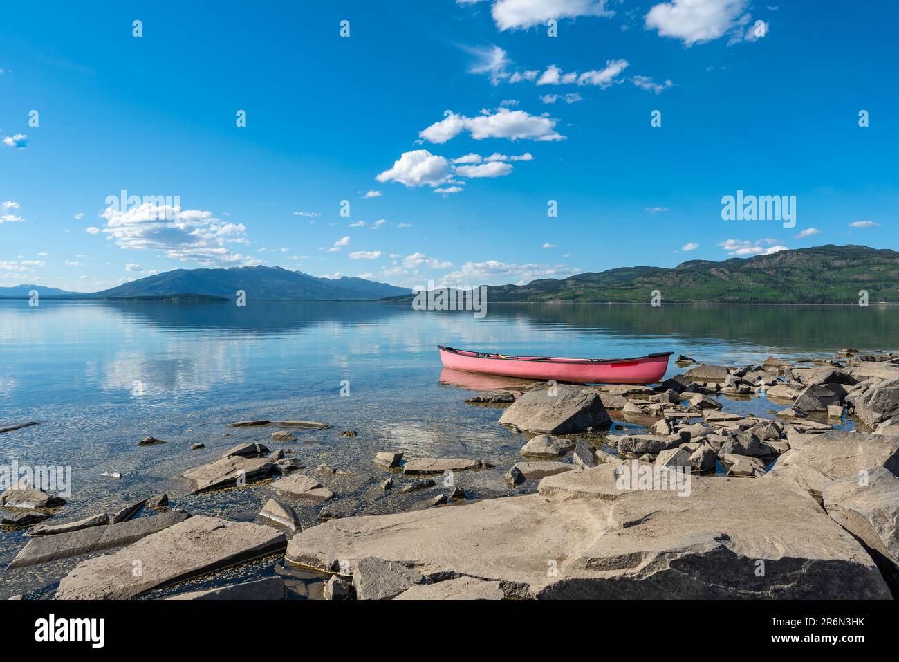 Red canoe sitting aside a rocky lake side on a beautiful blue sky summer day in northern Canada with mountains reflecting in the calm water below. Stock Photo