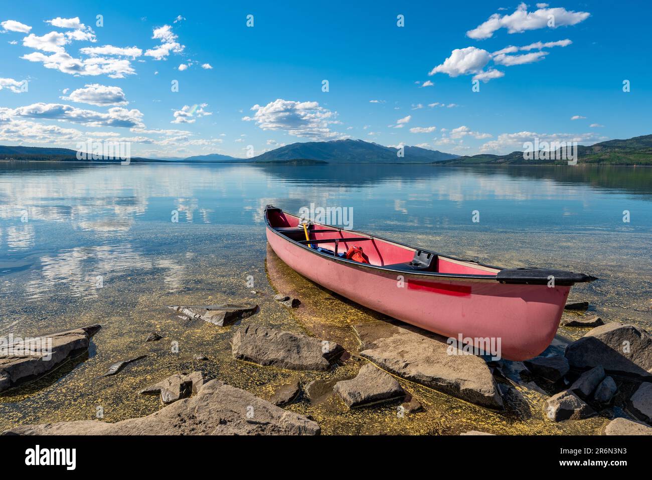 Red canoe sitting aside a rocky lake side on a beautiful blue sky summer day in northern Canada with mountains reflecting in the calm water below. Stock Photo