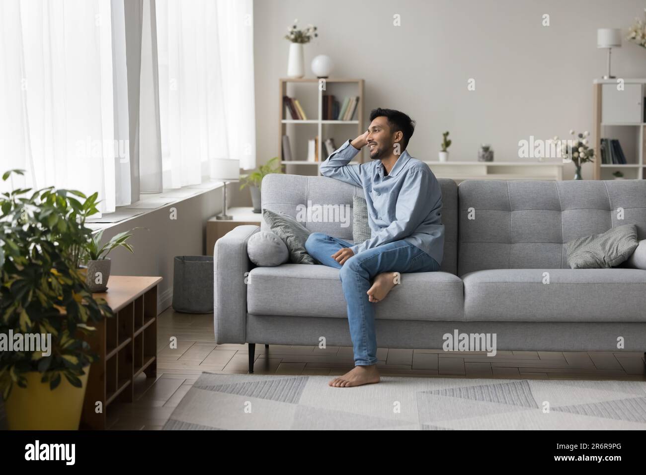 Positive dreamy relaxed young Indian guy sitting on comfortable couch Stock Photo