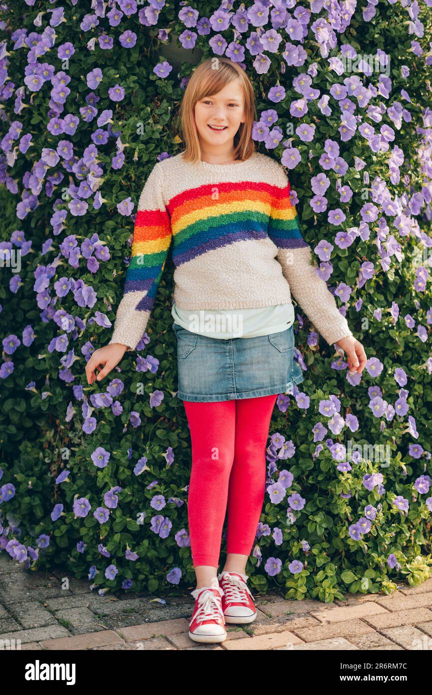 Outdoor portrait of pretty little girl wearing rainbow pullover and denim skirt, posing next to Morning Glory flowers wall Stock Photo