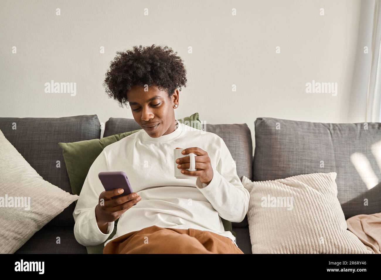 Relaxed calm gen z African American teen sitting on couch using phone. Stock Photo