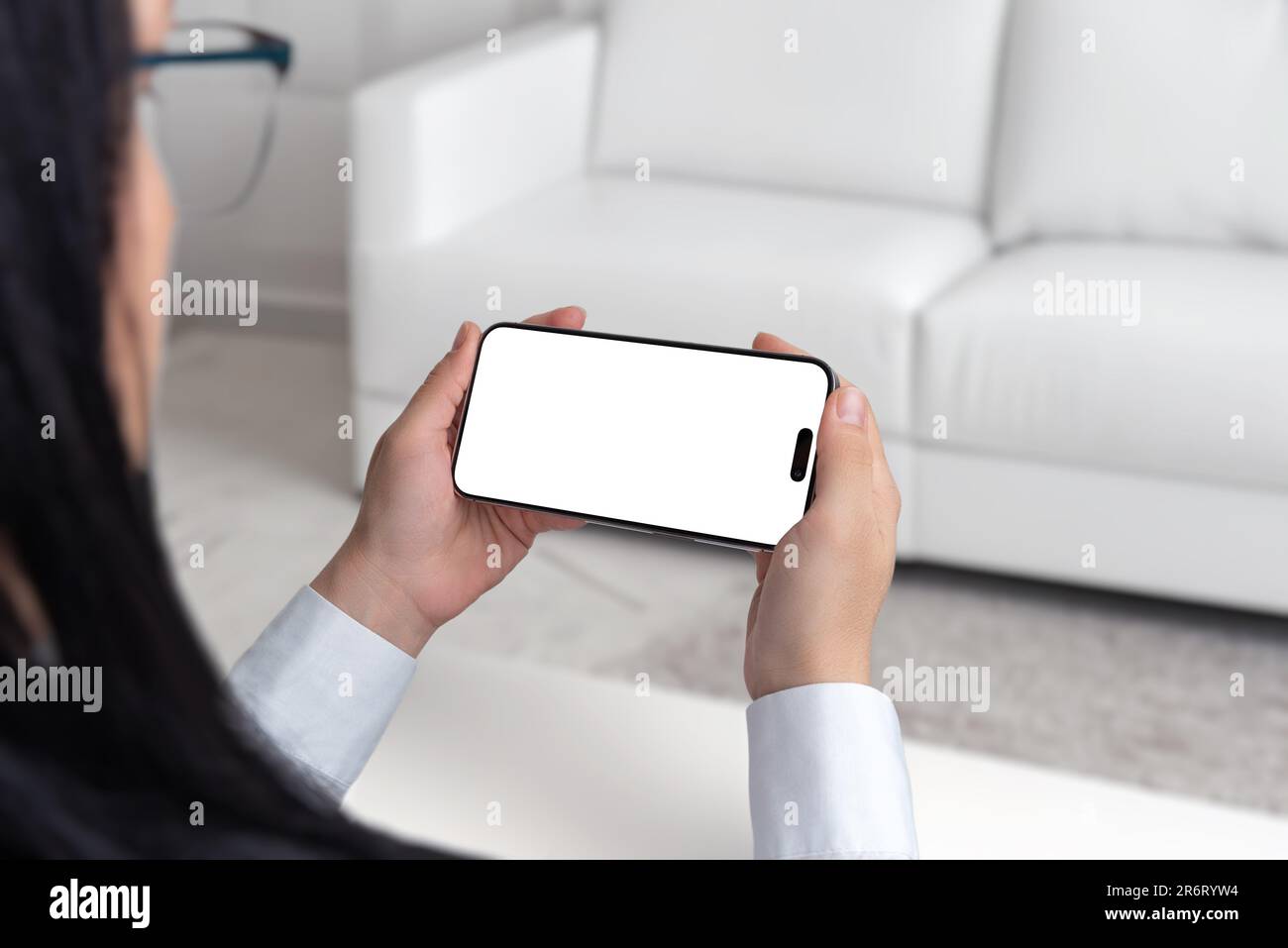 Young woman is sitting on a white couch, holding and watching smartphone in horizontal position. Isolated screen in white for mockup, app presentation Stock Photo