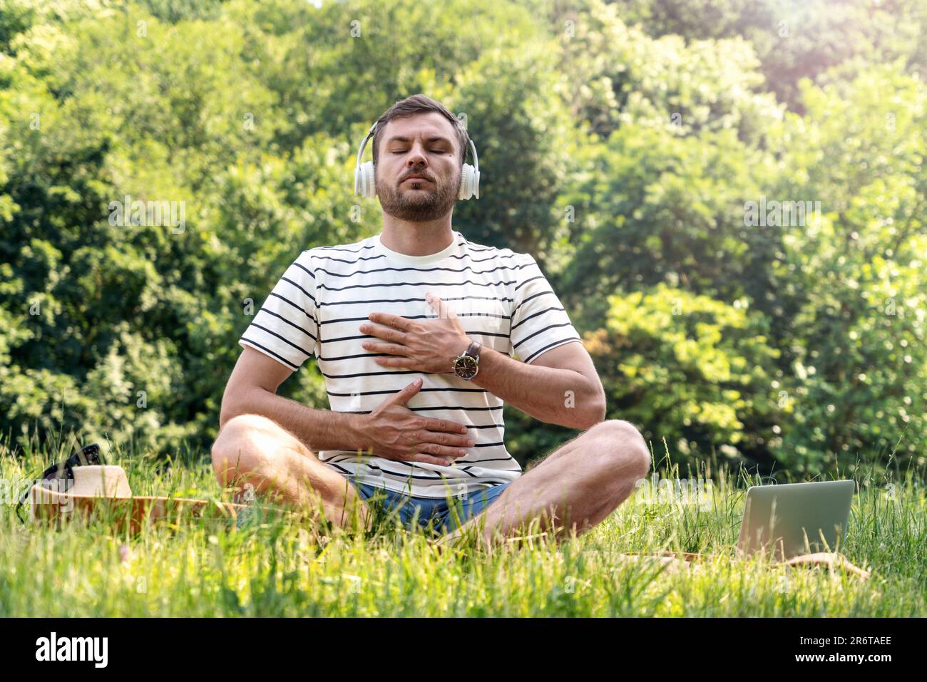 Man wearing headphones doing yoga breathing exercise in half lotus position in the park, listening music for meditation. Stock Photo
