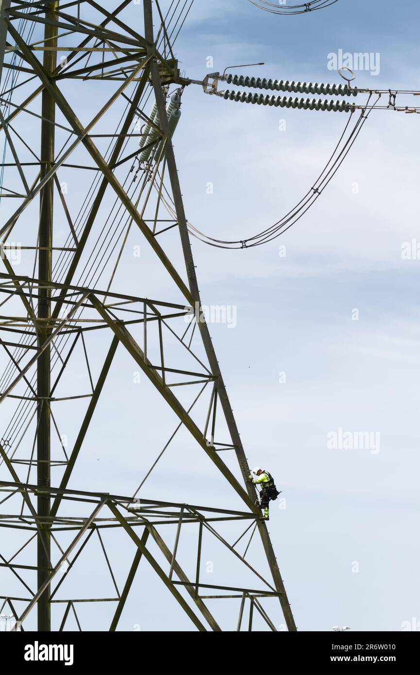 Electricity Worker In High Vis Jacket And Hard Hat Climbing Up A Metal Electricity Pylon To Repair IT, England UK Stock Photo