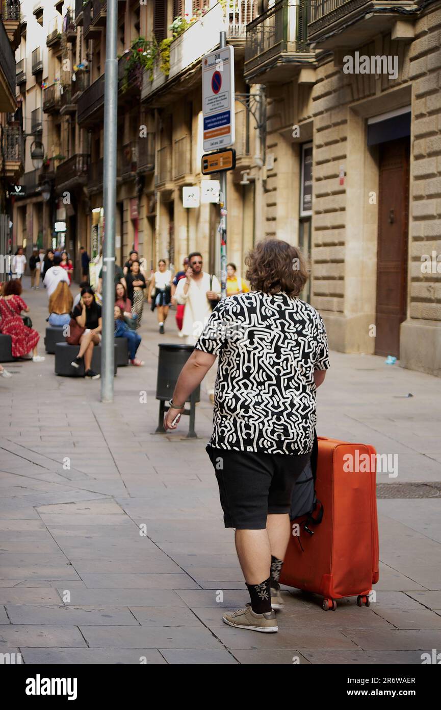 Barcelona - June 11, 2023: A cheerful unrecognizable tourist with a red suitcase explores the lively city, surrounded by colorful buildings. Experienc Stock Photo