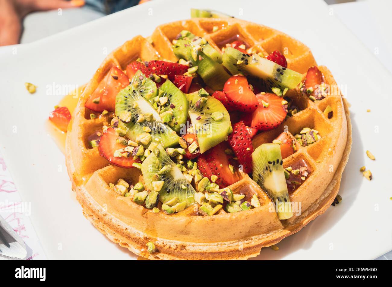 Large waffle with fresh strawberries, kiwi slices, pistachio pieces and honey on white plate Stock Photo