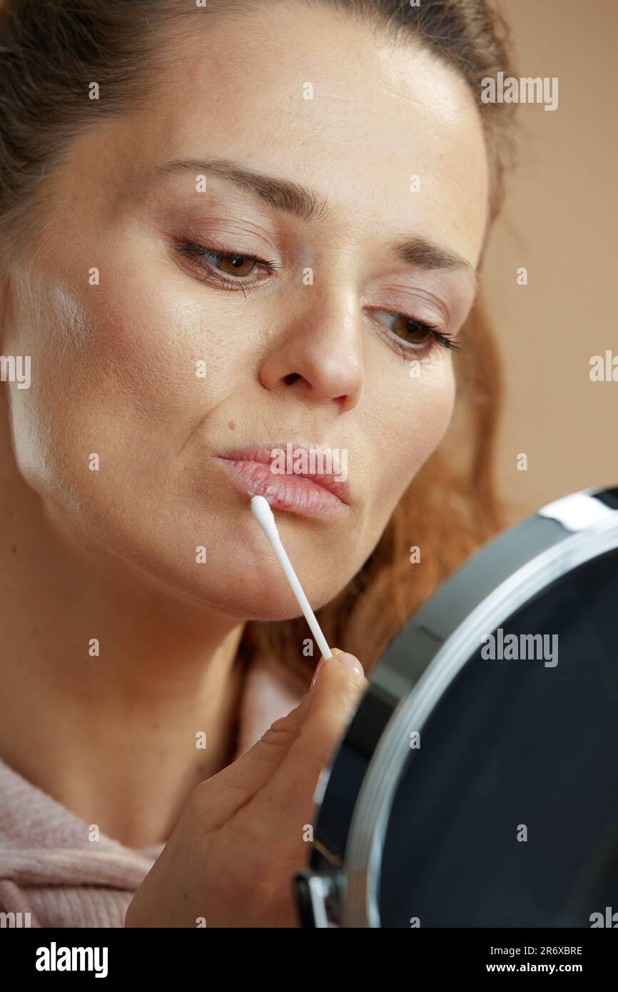 woman with herpes on lips and mirror applying ointment using cotton swab isolated on beige