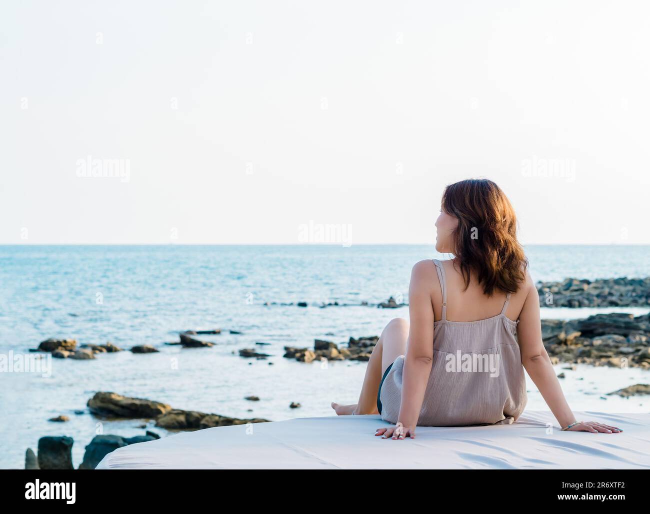 Back of Asian woman in casual dress sit in front of the beach in summer vibes. Lonely female relaxing smile at the sea and looking far away, seascape Stock Photo