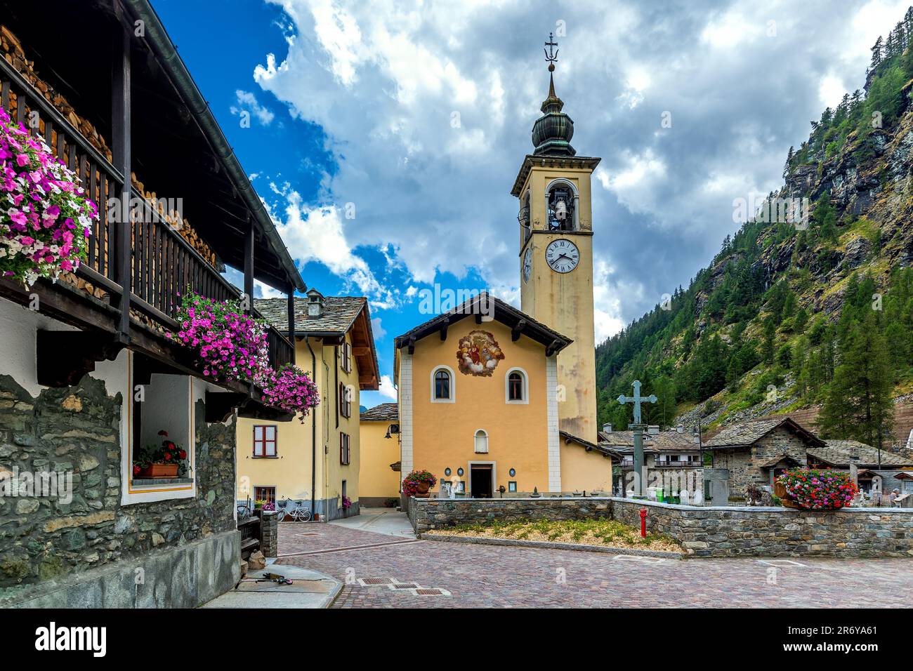 Church with big clock on the bell tower and houses decorated with flowers in small alpine town of Gressoney la Trinite in Italy. Stock Photo