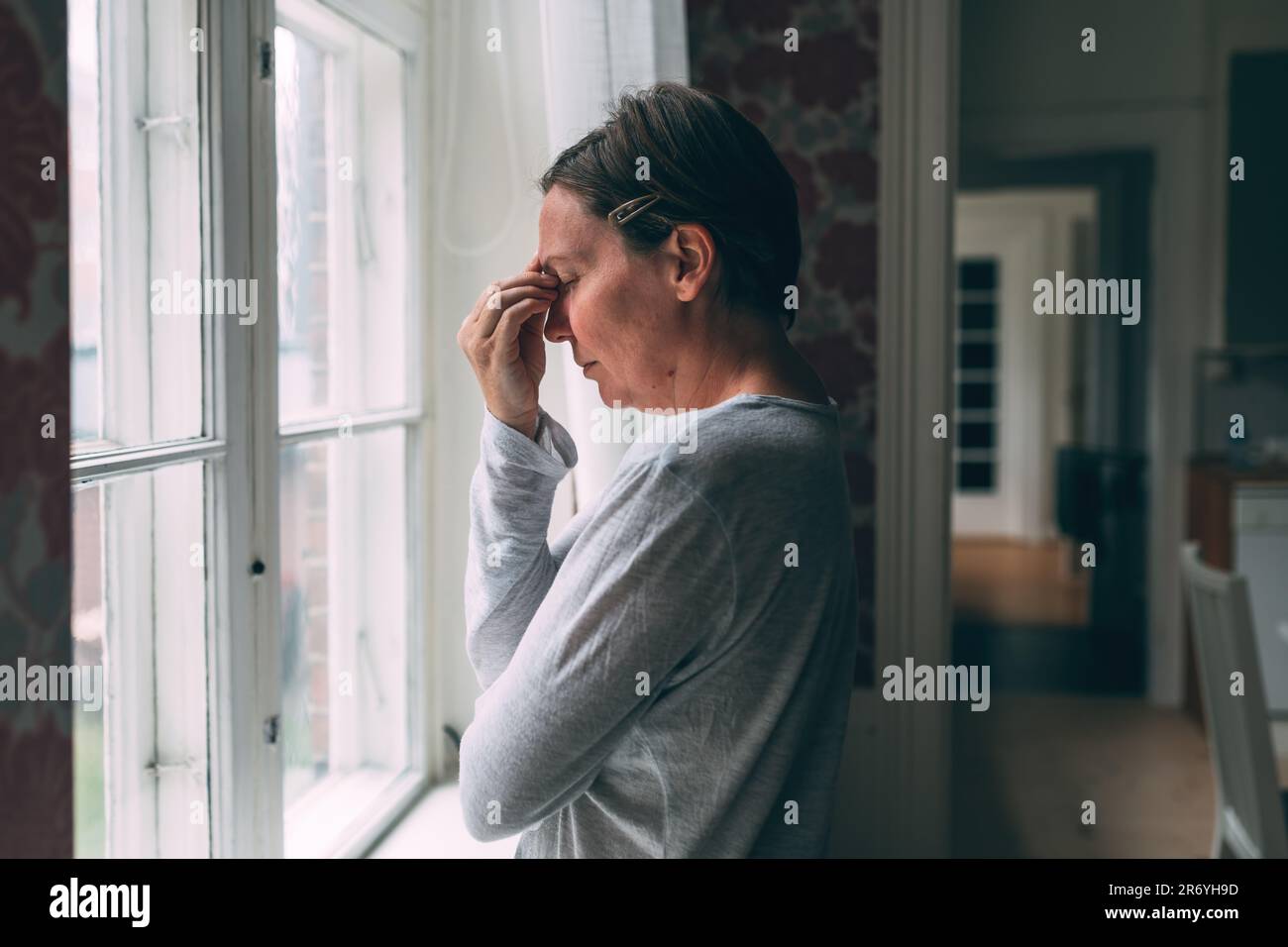 Mid-adult caucasian woman with severe headache standing by the living room window in morning and pressing the forehead with fingers, selective focus Stock Photo