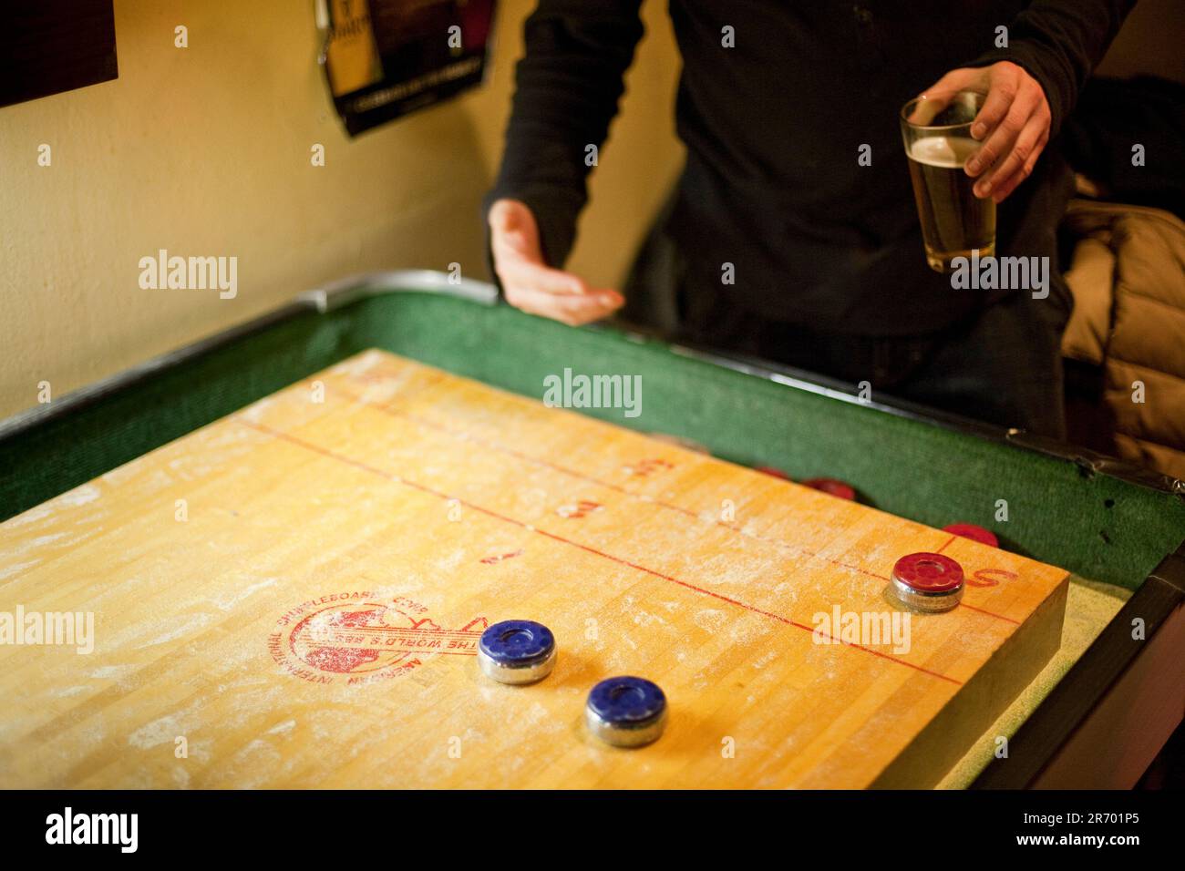 A man leans over a vintage wooden shuffleboard table while holding a