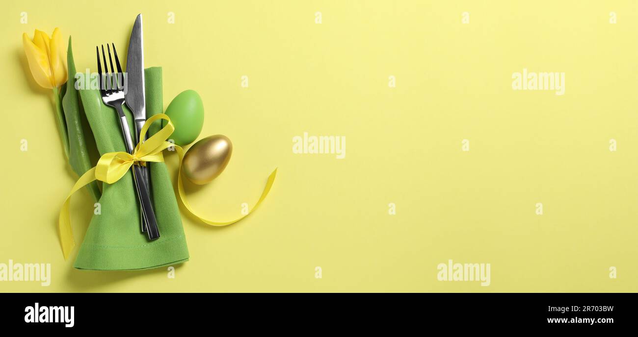 Set of cutlery, Easter eggs and tulip on yellow table, top view. Banner ...